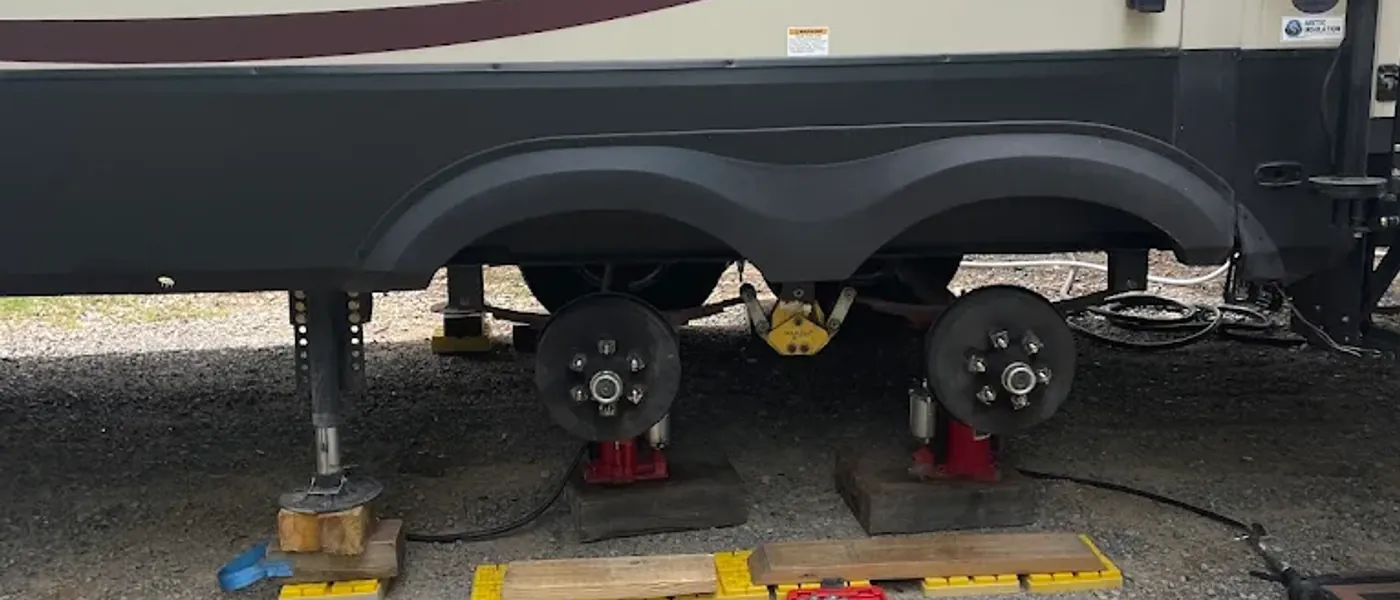 Travel trailer raised on jack stands with both wheels removed, bare hubs exposed, leveling blocks and an impact wrench laid out on the ground
