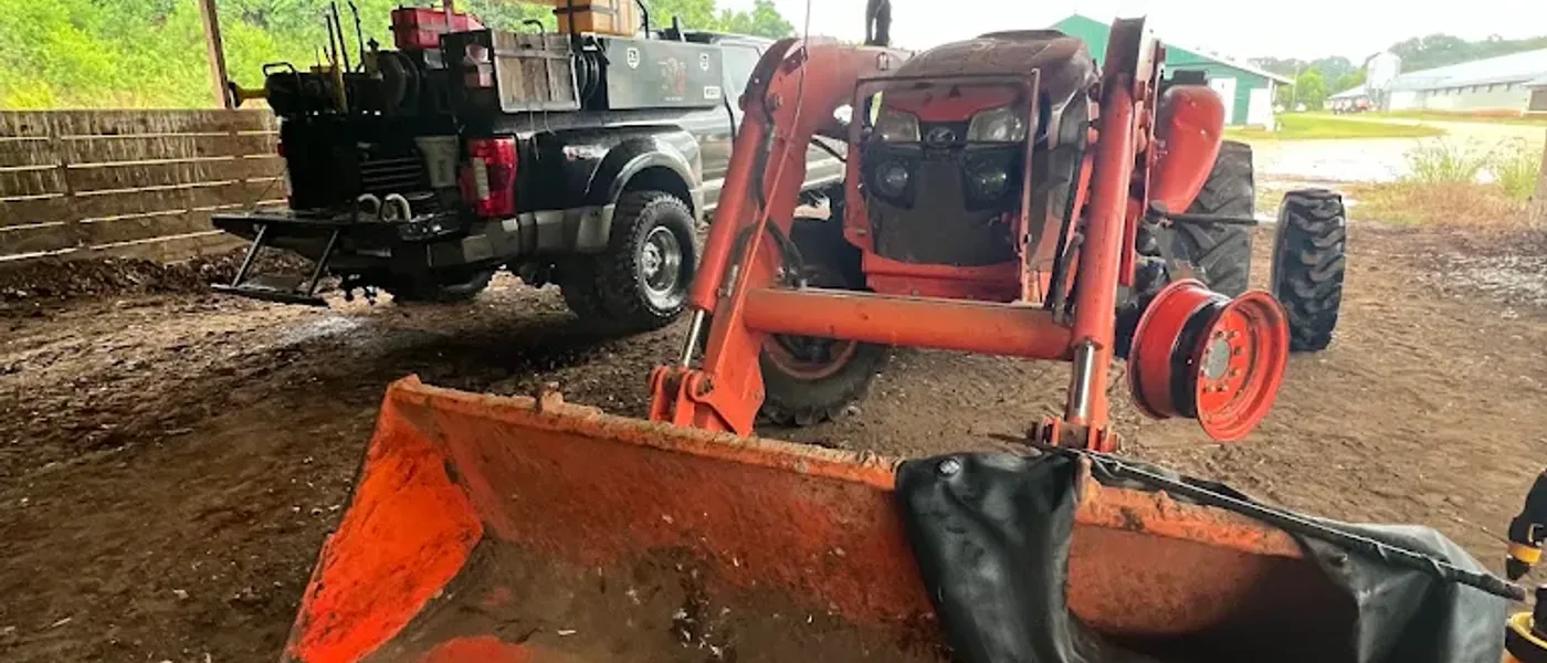 Orange front-loader tractor under a barn with a rear wheel removed and a new tube laid across the loader bucket, Breaux's Roadside service truck parked behind