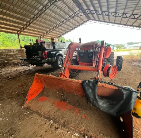 Orange front-loader tractor under a barn with a rear wheel removed and a new tube laid across the loader bucket, Breaux's Roadside service truck parked behind